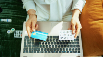 Upper view of woman with credit card and empty blister pack of pills buying pharmacy online using laptop at home in sunny day.