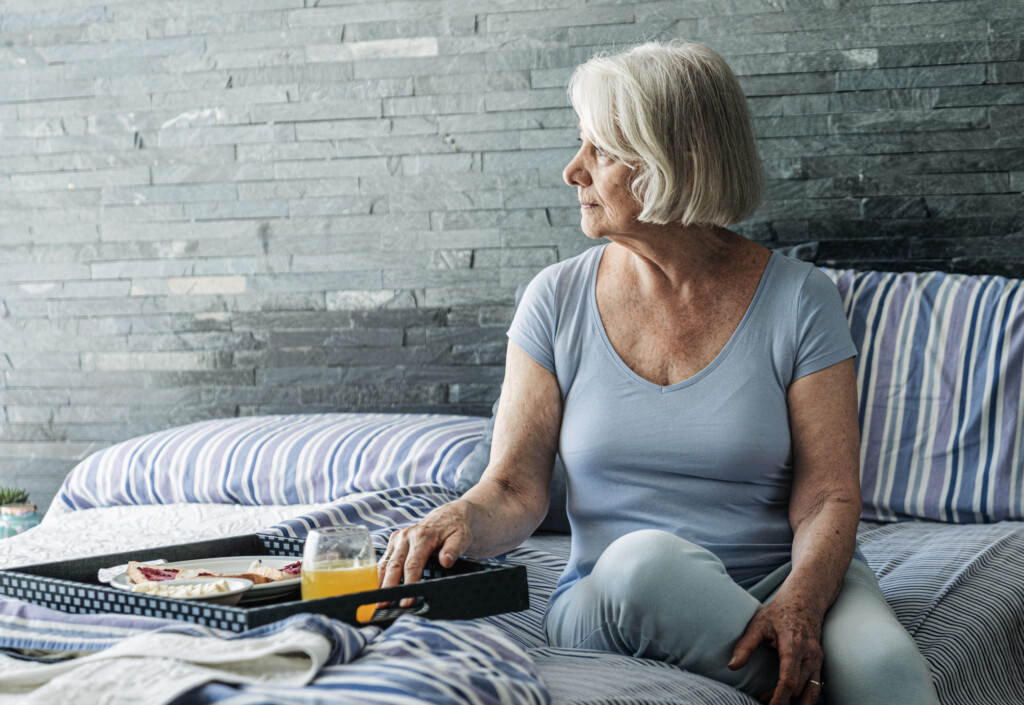 Contemplating elderly woman by food tray looking away. Sad senior female having short hair is sitting on bed. She is in casuals.