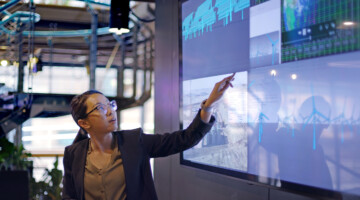 Woman pointing at several monitors with graphs.