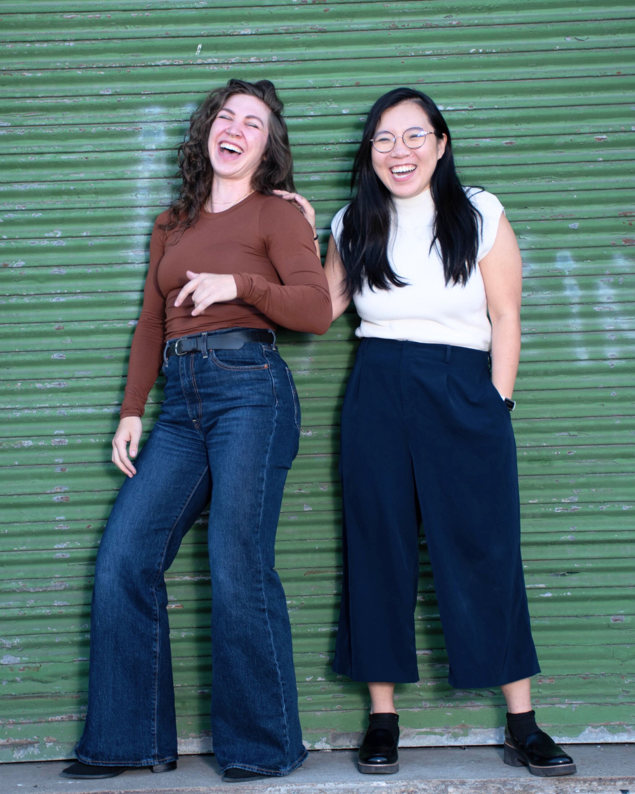 Two women standing in front of a green metal wall, laughing together.